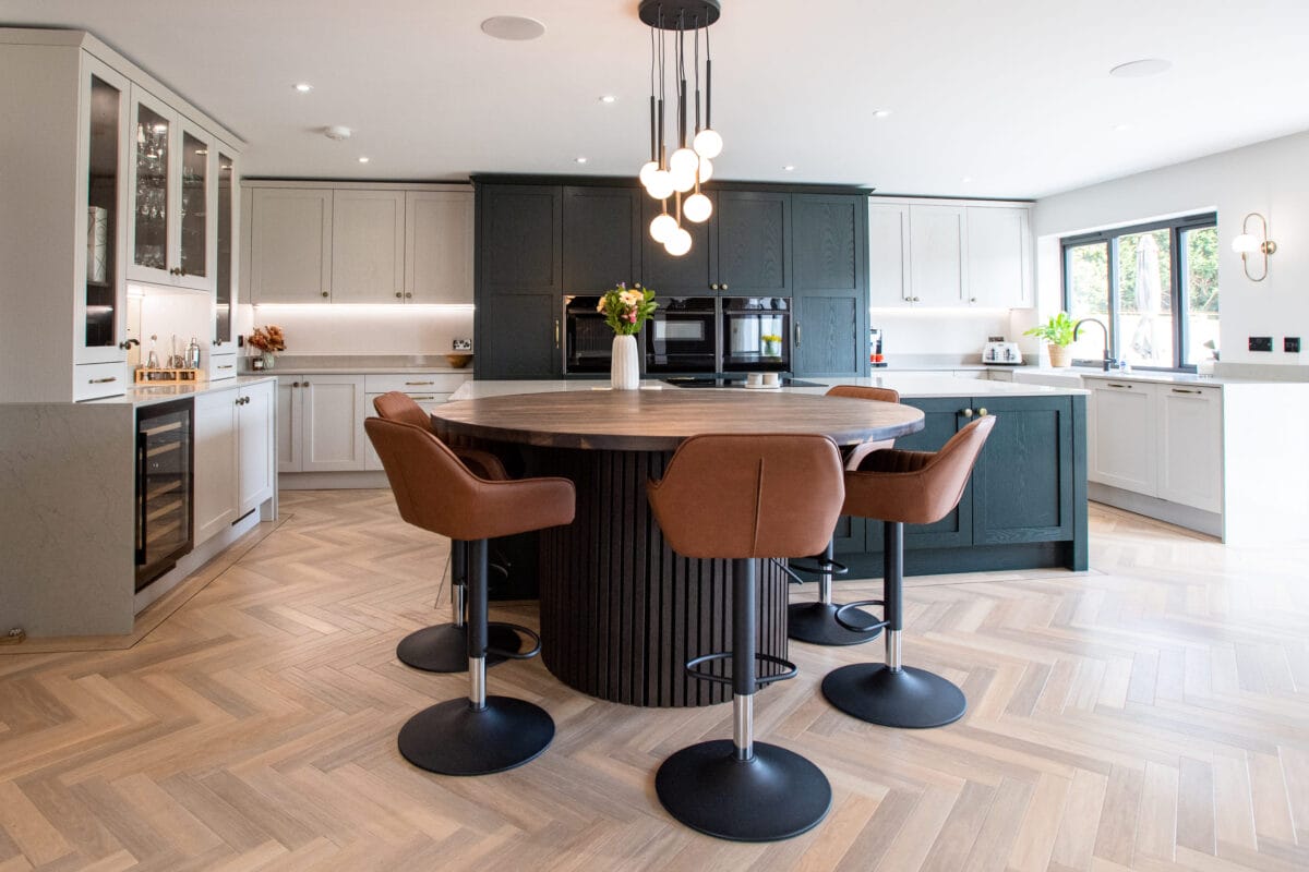 Two-tone shaker kitchen with dark green cabinetry, round oak island, leather bar stools and herringbone floor - Kitchen Living, Hampshire