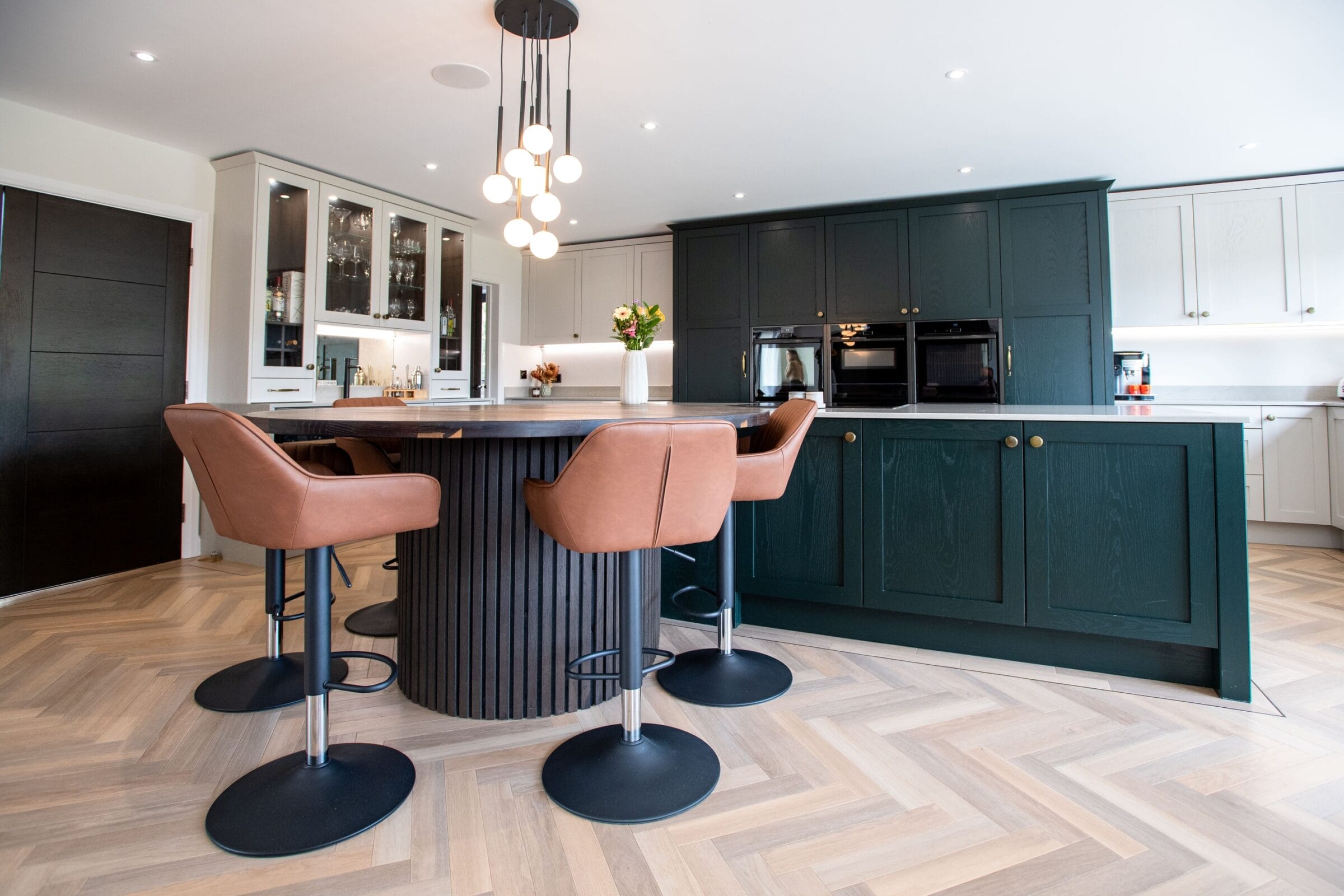 Dark green Kitchen Living kitchen with curved island, pink bar stools, herringbone flooring and modern pendant lighting