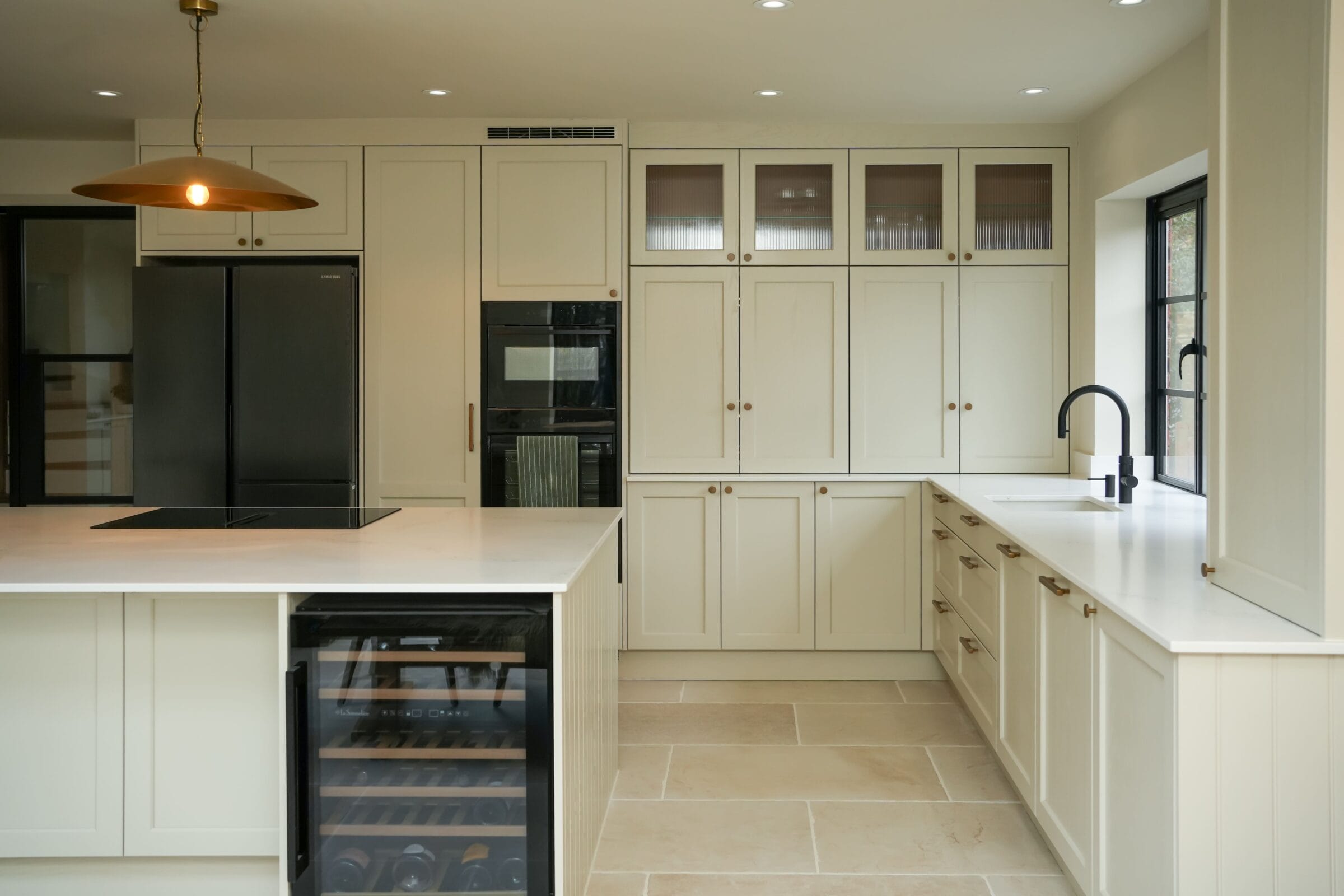 Cream shaker kitchen with integrated wine cooler, tall larder cabinets, quartz worktops and limestone tile flooring by Kitchen Living
