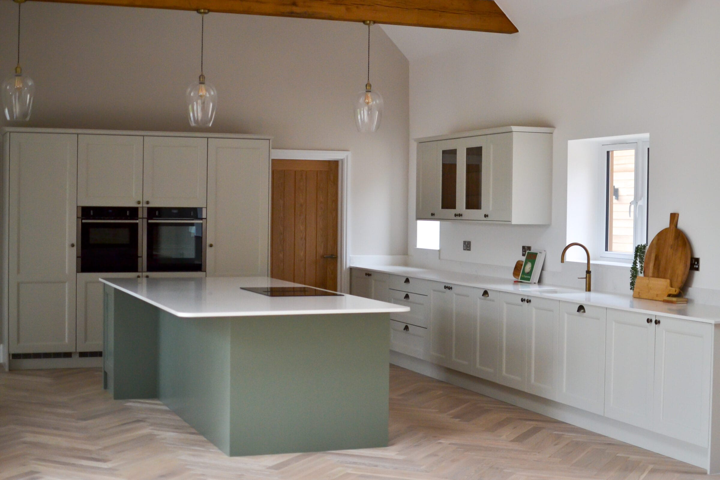 Sage green and off-white shaker kitchen with central island, quartz worktop and herringbone wood floor in Hampshire barn conversion by Kitchen Living